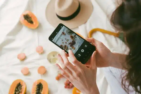 Um aparelho celular nas mãos de uma mulher editando a foto de uma mesa com frutas e um chapéu